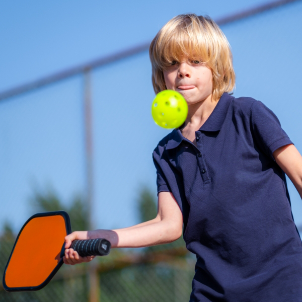 Young child with blond hair playing pickleball outdoors on wide pickleball courts, skillfully hitting a yellow ball with a paddle, wearing a navy shirt, with a fence and trees in the background.