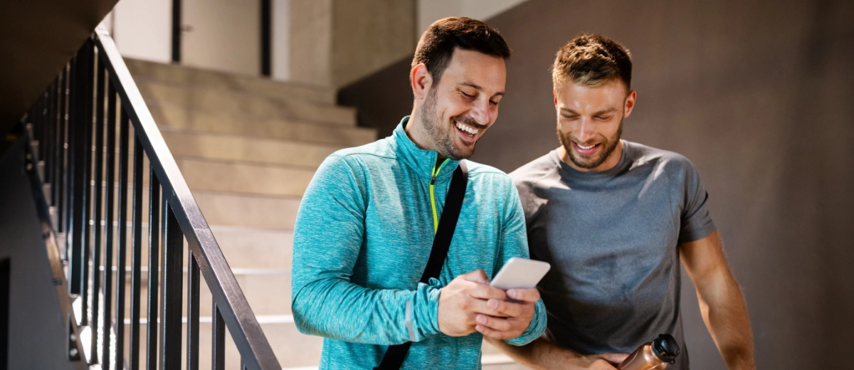 Two men stand on a staircase at the pickleball center, one wearing a blue sweatshirt and holding a phone, smiling. The other, in a gray shirt, holds a water bottle, looking at the phone.