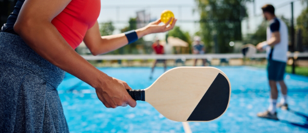 Close-up of a person holding a pickleball paddle, about to serve a yellow ball. Two players are seen in the background at an outdoor court, making the most of their time at this versatile pickleball facility.