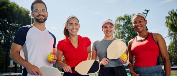 Four people holding paddles and pickleballs stand together on an outdoor court, smiling at the camera.