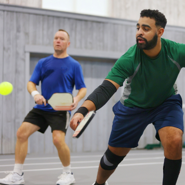 Two men engage in a lively pickleball match on the indoor pickleball court. One is readying his paddle to serve, while the other keenly observes. Both are dressed in casual sports attire, perfectly suited for the energetic atmosphere of this dedicated pickleball center.