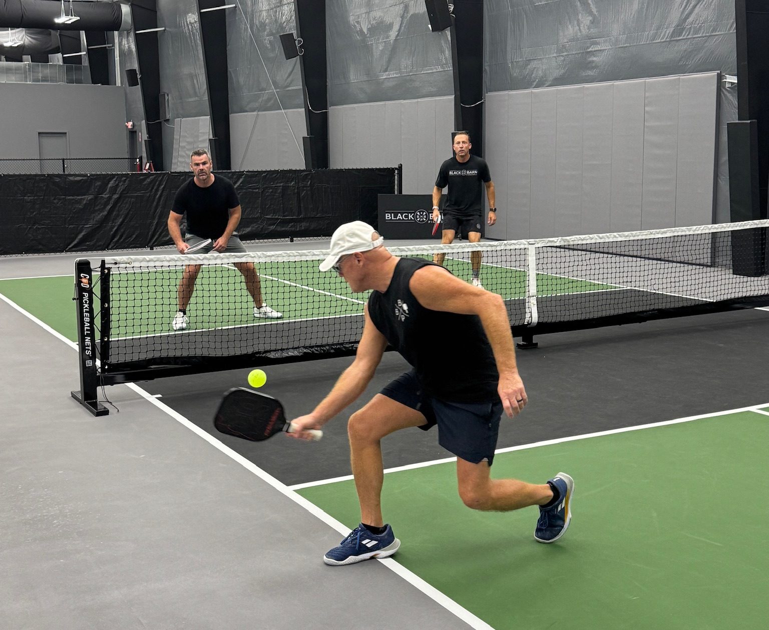 Three men play pickleball on one of the Best Pickleball Courts indoors; one man in front reaches to hit the ball with his paddle while the other two stand near the net on the opposite side.