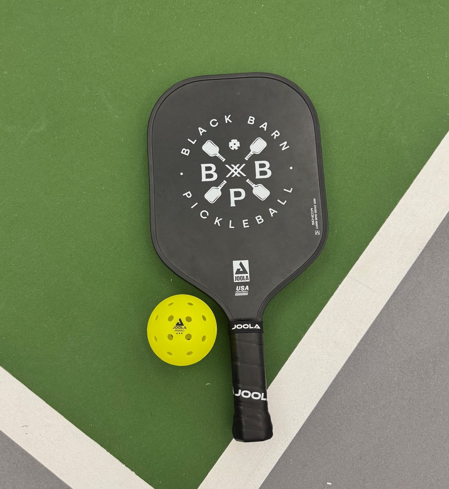 A black pickleball paddle labeled "Black Barn Pickleball" and a yellow pickleball rest on one of the best pickleball courts, featuring a green and gray court surface.