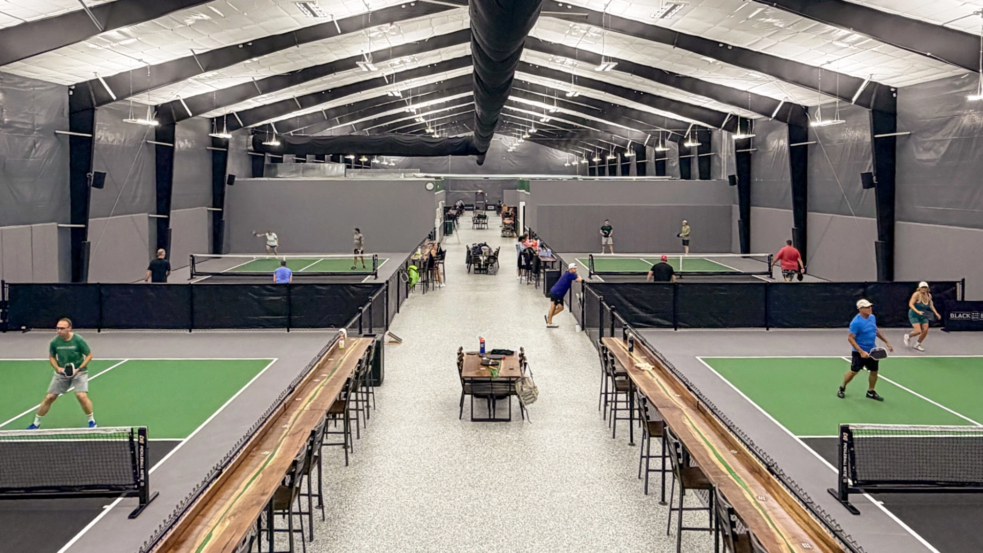 An indoor sports facility with several pickleball courts, where multiple groups of people are playing games under a high, industrial ceiling.