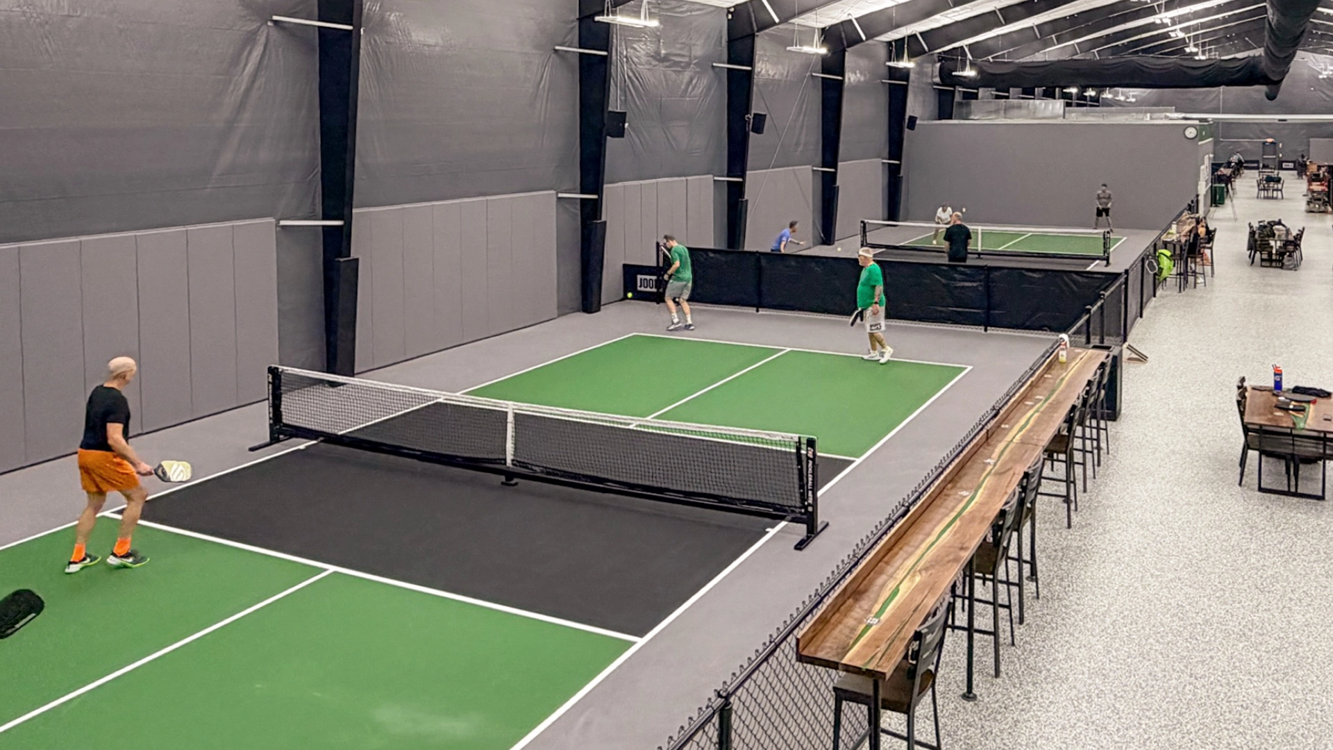 Indoor facility with people playing pickleball on multiple courts, high ceilings, and seating areas with tables and chairs along one side.
