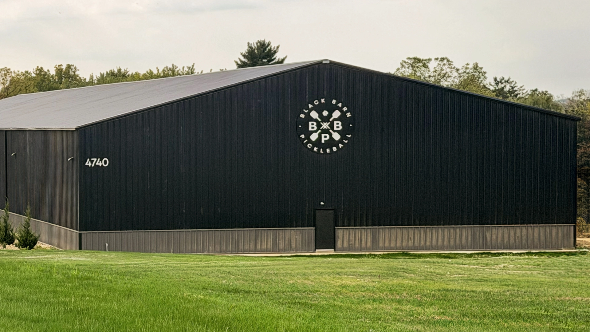A large black building with the address 4740 and a circular "Black Barn Pickleball" logo on the front, surrounded by grass and trees.